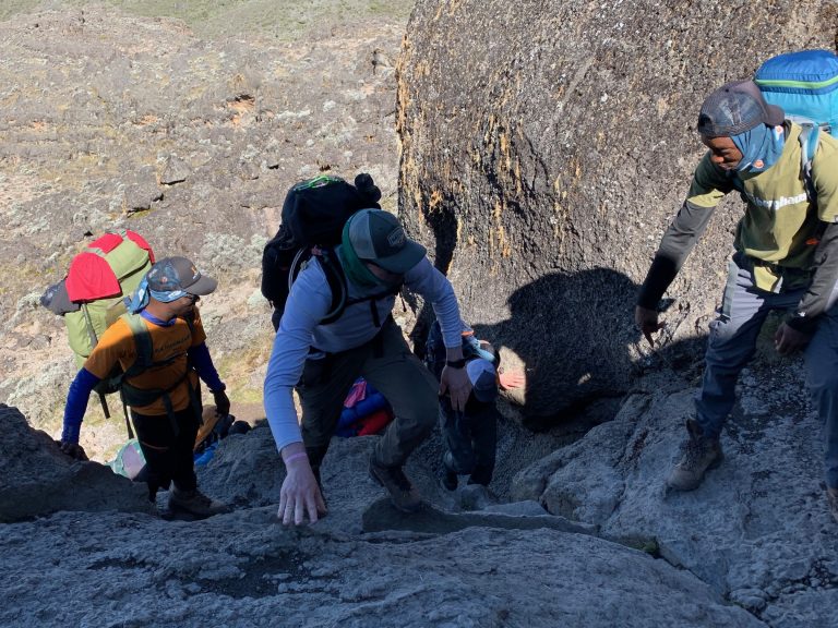 Two porters and a tourist trekking on a rocky path during a Kilimanjaro climb via Rongai Route